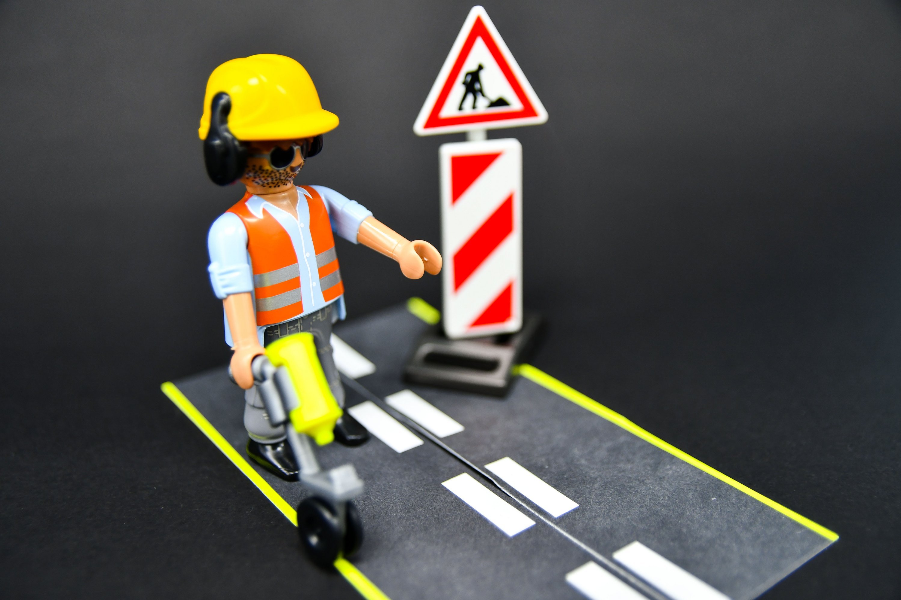 Eine Spielzeugfigur in Bauarbeiterkleidung steht auf einem kleinen Stück Spielstraße. Im Hintergrund ist eine Absperrung mit Baustellenschild aufgebaut. 