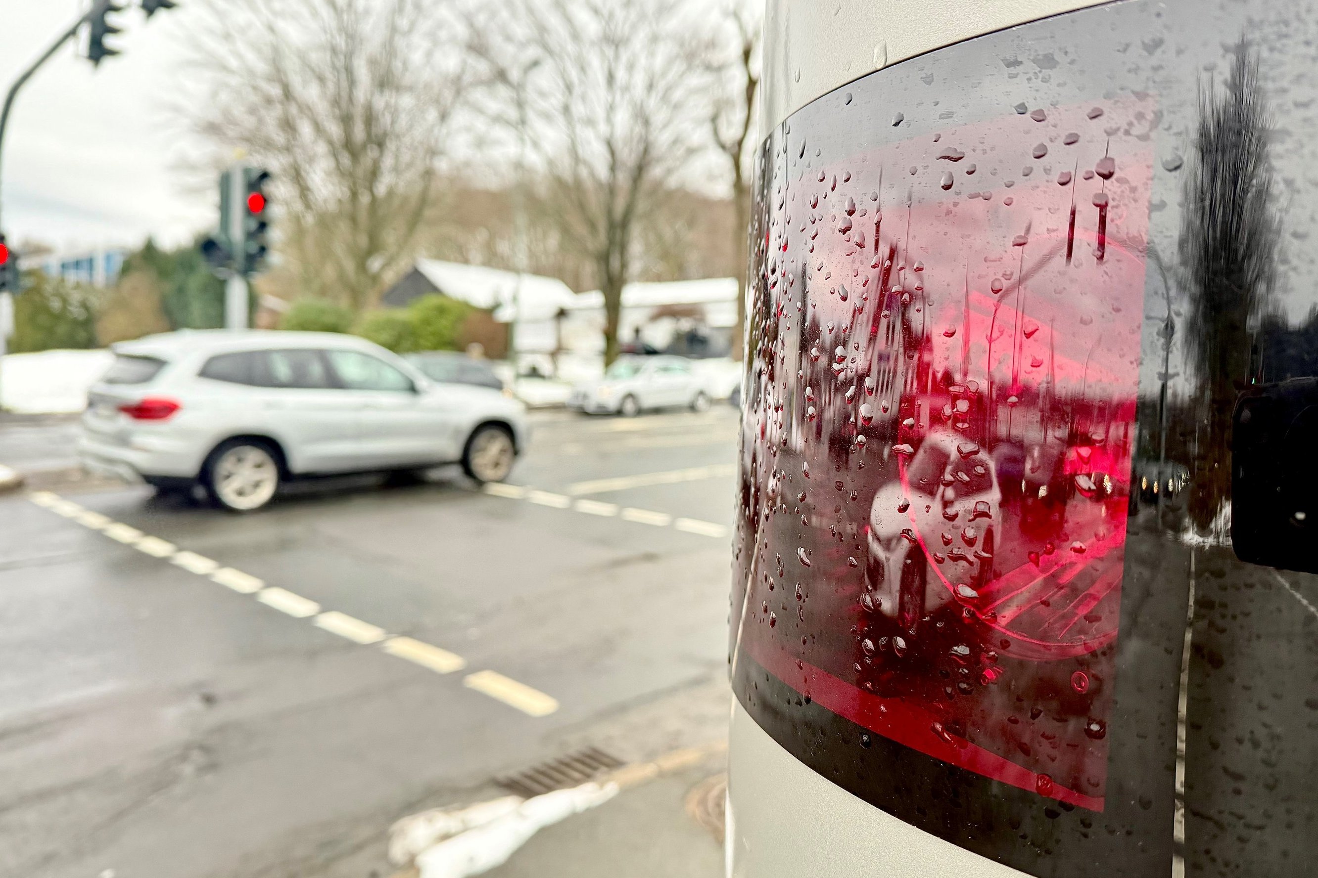 Im Vordergrund ist ein Lasermessgerät einer Blitzersäule zu sehen, das hinter einer roten Scheibe sitzt. Im Hintergrund fährt auf der Straße ein Auto vorbei..