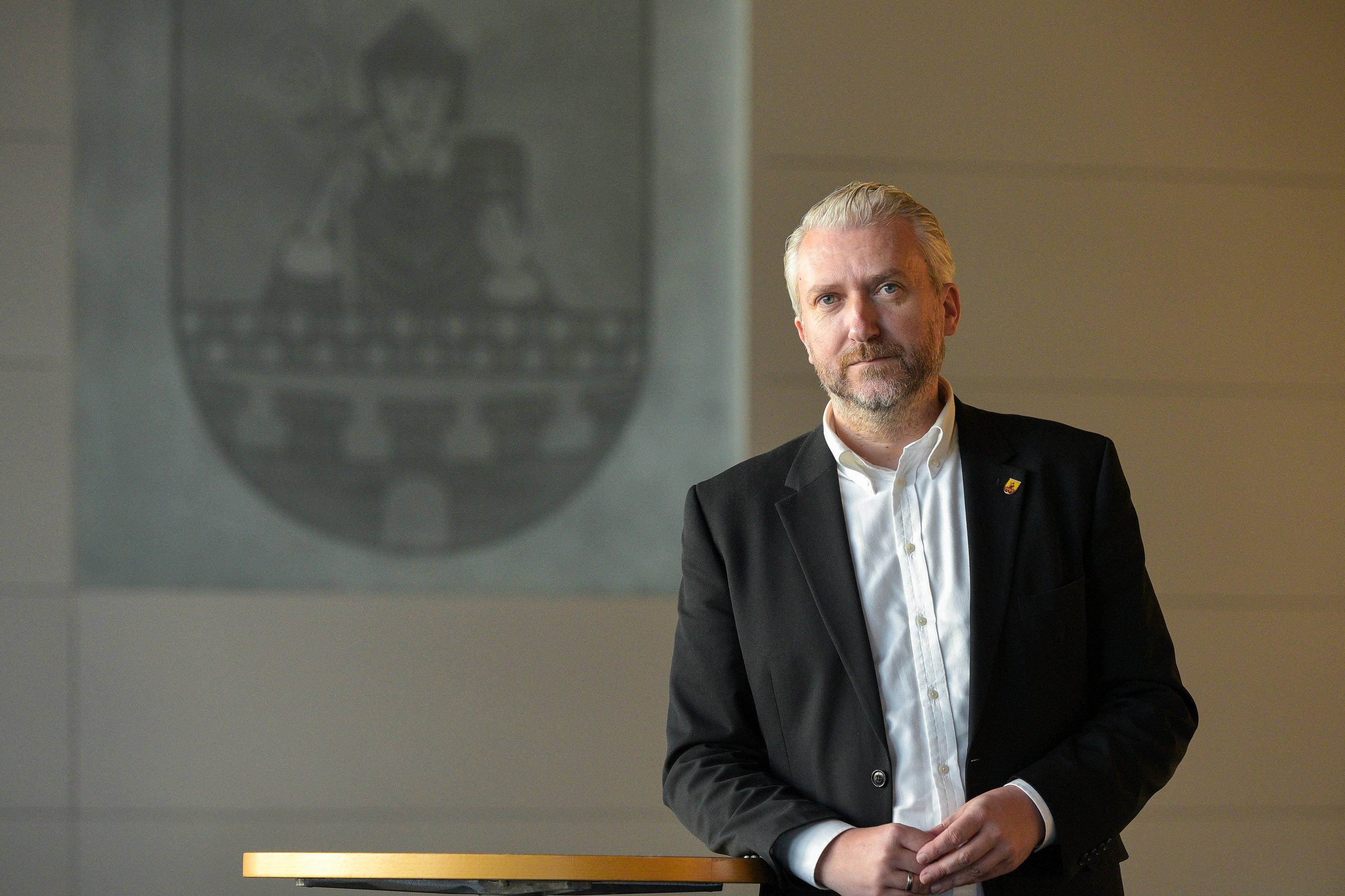 Bürgermeister Sebastian Wagemeyer steht im Ratssaal an einem Tisch. Im Hintergrund ist das Lüdenscheider Stadtwappen zu sehen.