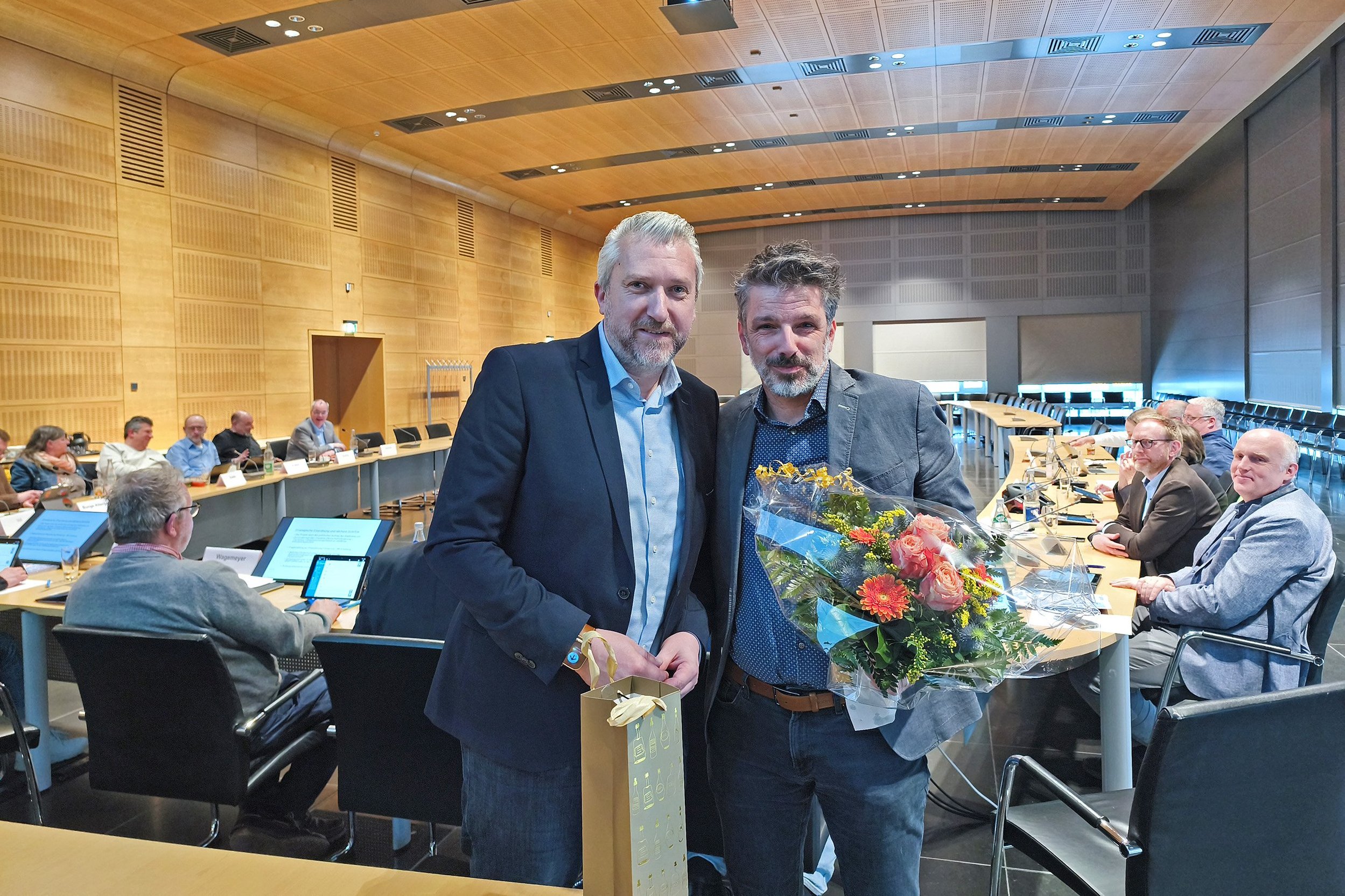Bürgermeister Sebastian Wagemeyer und der scheidende Leiter des Fachbereichs Klima und Umwelt, Marcus Müller, stehen zusammen im Ratssaal. Im Hintergrund sind Mitglieder des Hauptausschusses zu sehen.