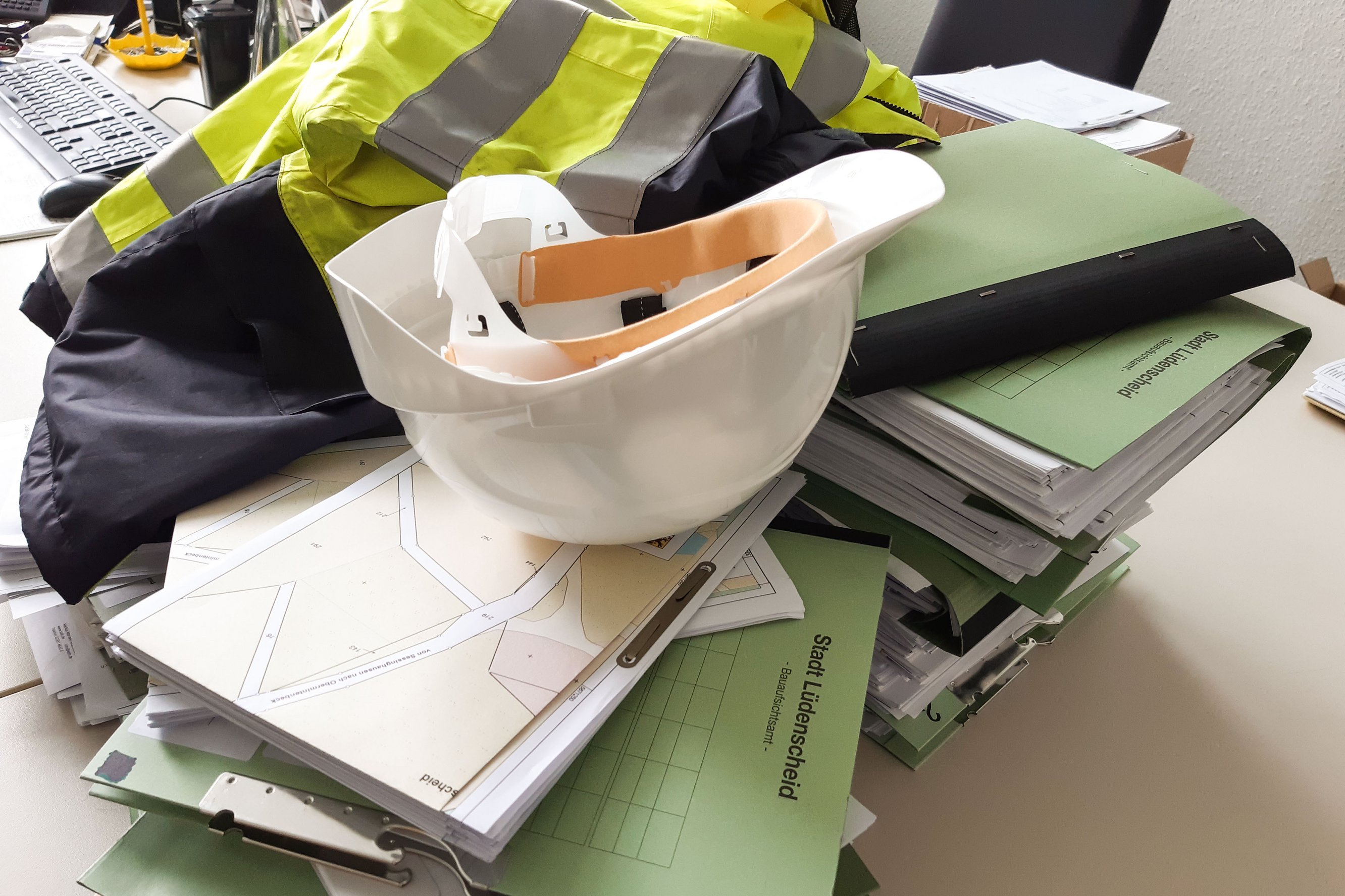 Akten, ein Baustellenhelm und eine Warnweste auf einem Schreibtisch