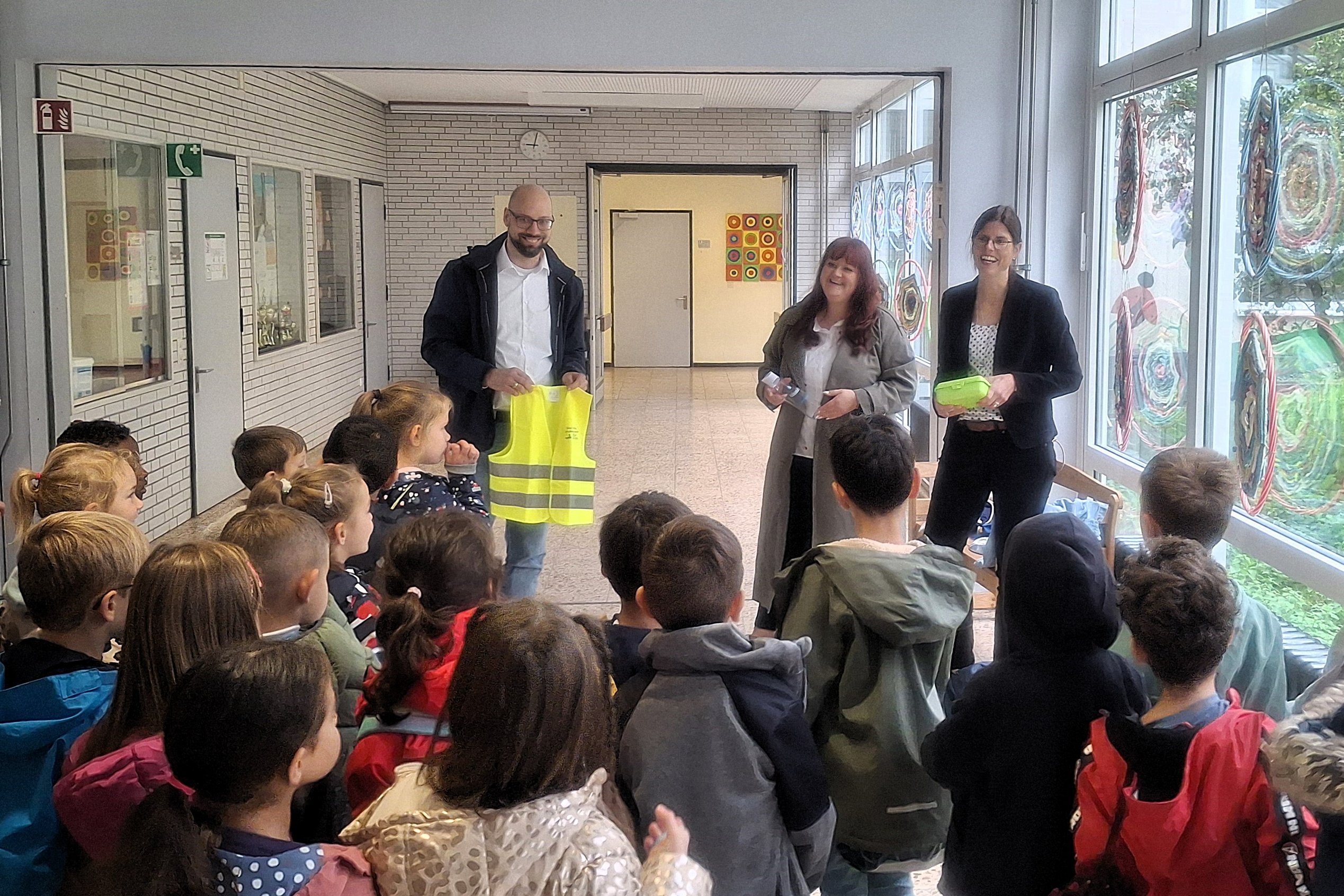 Drei Erwachsene verteilen Brotdosen, Trinkflaschen und Warnwesten an Erstklässlerinnen und Erstklässler.