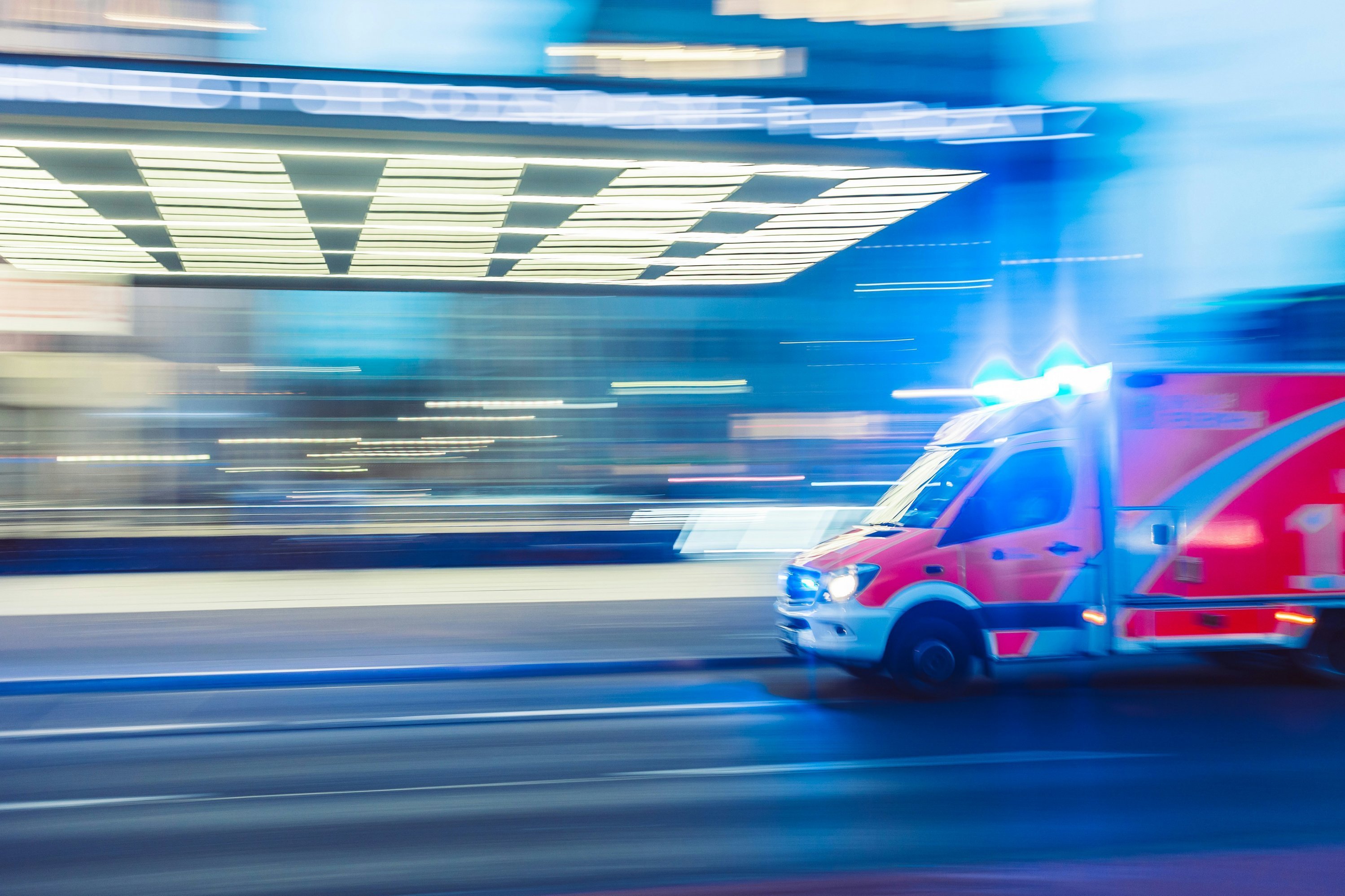 Ein Rettungsfahrzeug fährt mit eingeschaltetem Blaulicht über eine Straße. Das Bild ist bewusst unscharf gehalten, um Geschwindigkeit zu assoziieren. 