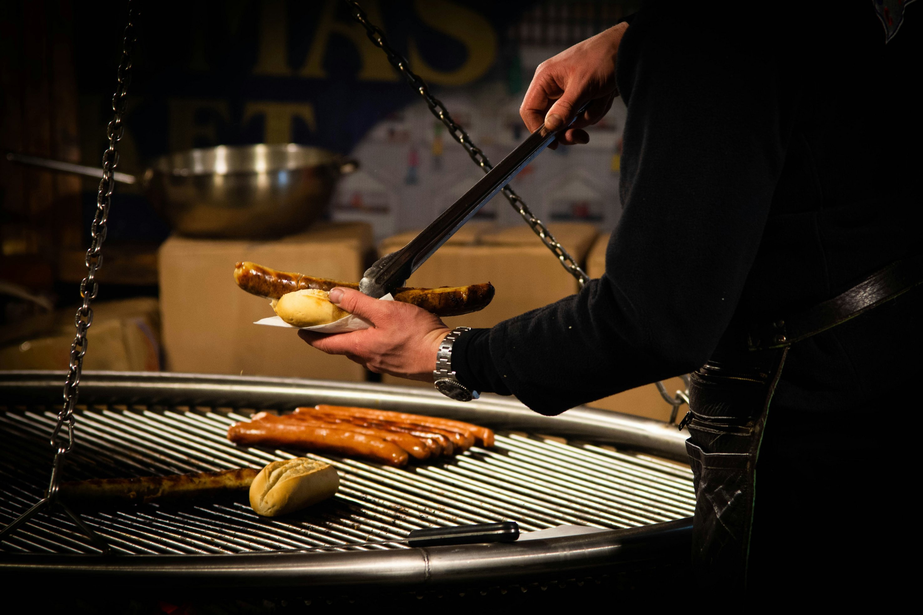 Eine Person steht am Grill und legt mit einer Zange eine Bratwurst in ein Brötchen.